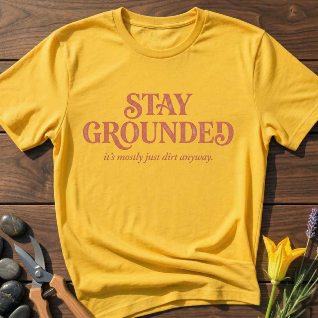 Yellow t-shirt with 'Stay Grounded' text on a wooden surface with gardening tools and flowers.