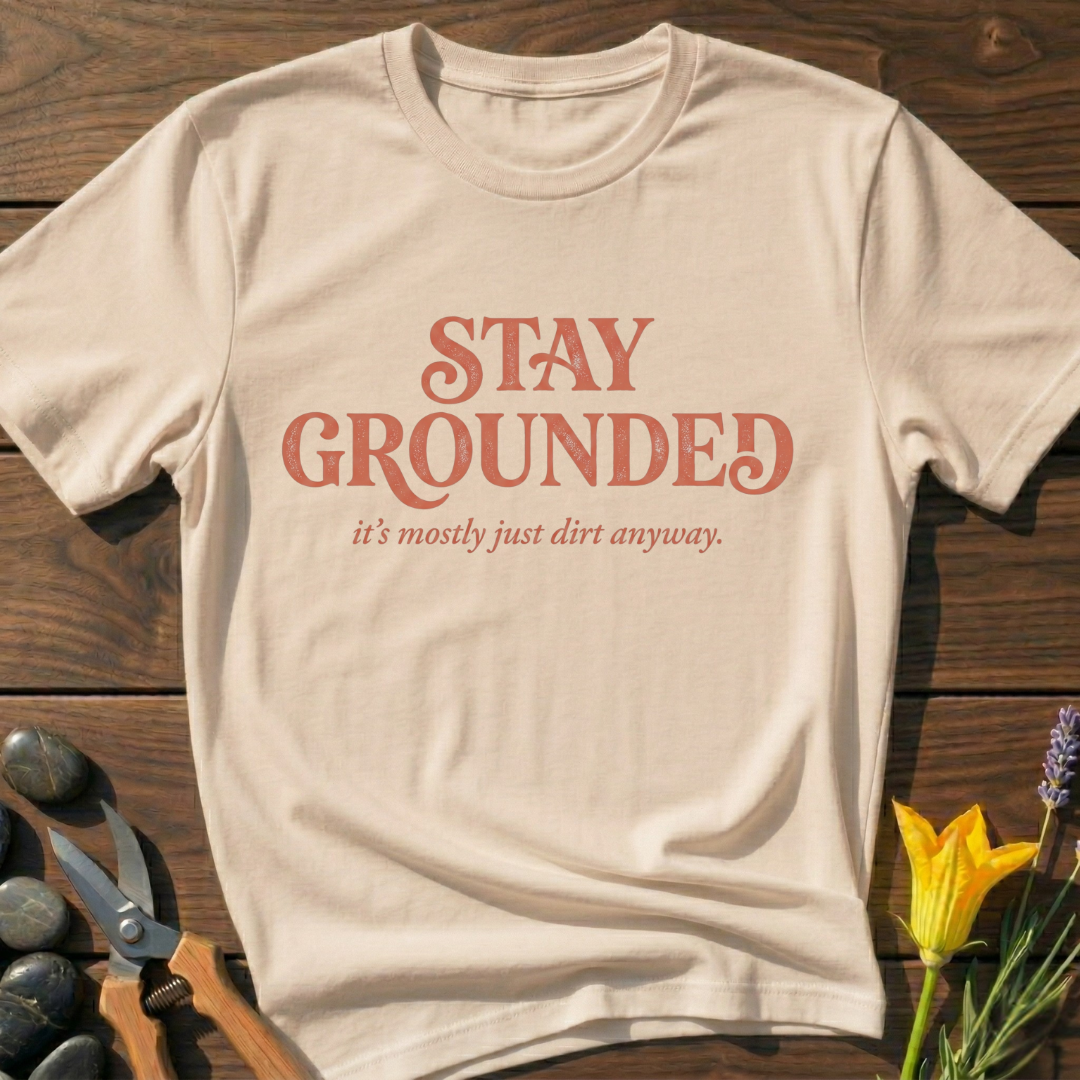 Beige t-shirt with 'Stay Grounded' text on a wooden surface with gardening tools and flowers.