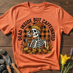 Orange t-shirt with skeleton graphic and text 'Dead Inside But Caffeinated' on a wooden surface.